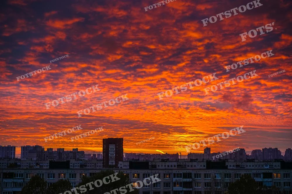 Это фотография городского пейзажа на закате с захватывающим небом, заполненным облаками, окрашенными в яркие оттенки красного и оранжевого цветов. Силуэты многоэтажных жилых домов очерчены против фона заходящего солнца, что создаёт контраст между тёмными зданиями и пылающим небом. Такие кадры часто используются для иллюстрации красоты и драматизма городской жизни или могут быть использованы в различных публикациях для добавления атмосферности.