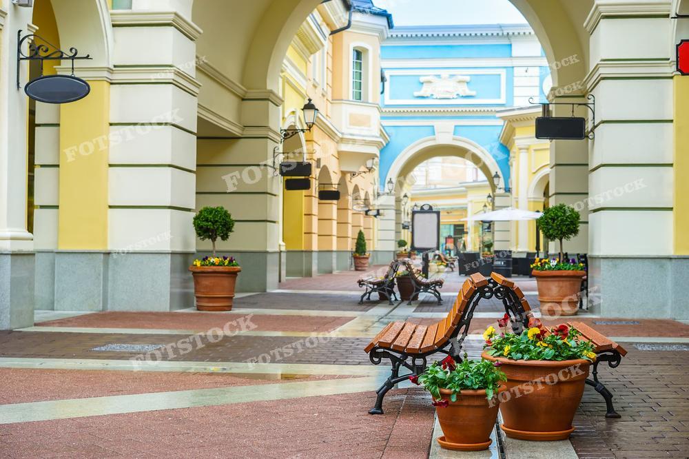 Фотография демонстрирует уютный городской дворик с архитектурой в классическом стиле. Яркие цветочные клумбы в керамических горшках украшают пространство, создавая чувство ухоженности и комфорта. Скамейки приглашают отдохнуть и насладиться атмосферой, а фонари добавляют изысканности. Такой вид может принадлежать историческому центру города или торговому комплексу, стилизованному под старину. Изображение подходит для туристических брошюр, материалов о градостроительстве, дизайне урбанистических пространств или как пример благоустройства городской среды.