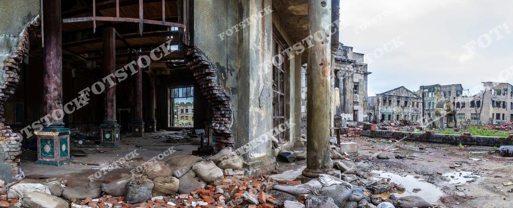 На фотографии изображены разрушенные колонны и пробитые стены здания, окруженные остатками кирпичей и мешков. На заднем плане видны руины других зданий, создавая мрачную и трагичную атмосферу. Снимок символизирует упадок и последствия исторических катастроф, оставивших свой отпечаток на архитектуре.