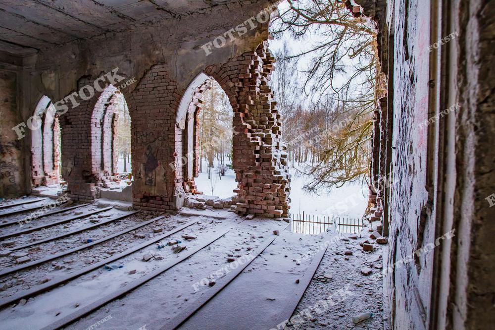 На фотографии изображены руины разрушенного кирпичного здания. Разбитые кирпичи лежат на полу, а в стенах видны дыры, отражающие запустение и заброшенность дома.