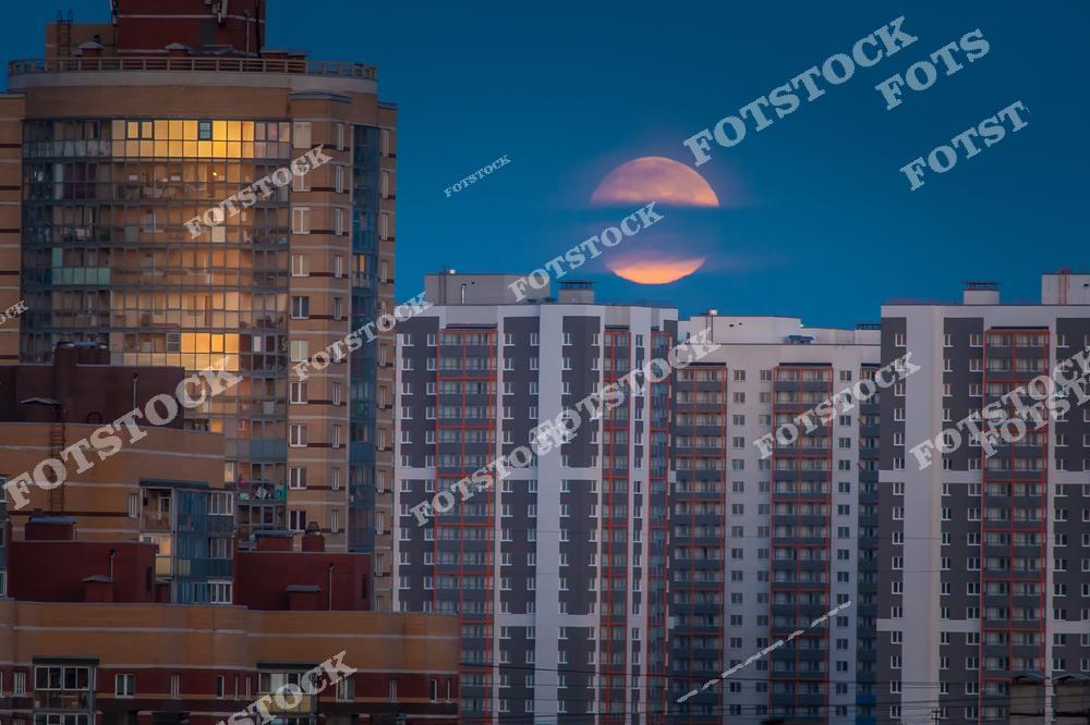 На фотографии изображен вечерний городской пейзаж с многоэтажными зданиями, где на фоне ясного ночного неба восходит Луна. Это изображение подчеркивает динамичность жизни в мегаполисе и концепцию урбанистики.