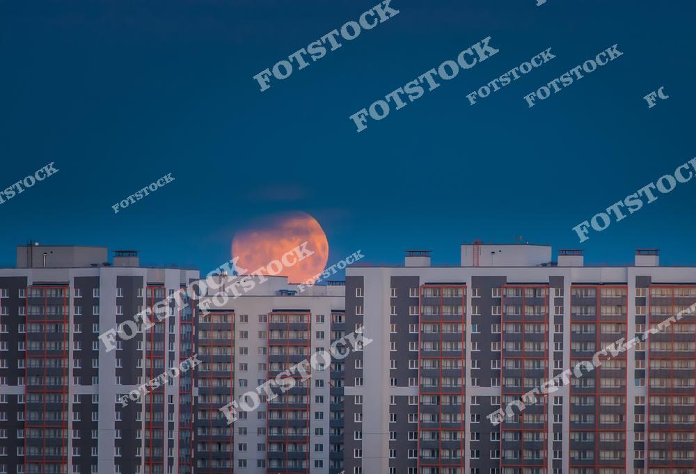 На фотографии изображены многоэтажные дома, над которыми поднимается яркая полная луна. Глубокое синее небо усиливает контраст между природной красотой и урбанистическим пейзажем. Снимок передает атмосферу вечера в современном городе.