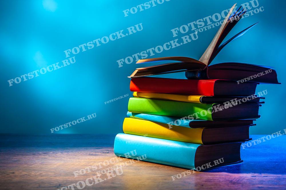 Фотография стопки книг с яркими разноцветными обложками, расположенных на деревянной поверхности. Верхняя книга открыта, её страницы развеваются. Синий фон создаёт спокойную и вдохновляющую атмосферу, подчёркивая насыщенные цвета обложек. Эта композиция идеально подходит для иллюстрации тем, связанных с чтением, образованием и литературой.