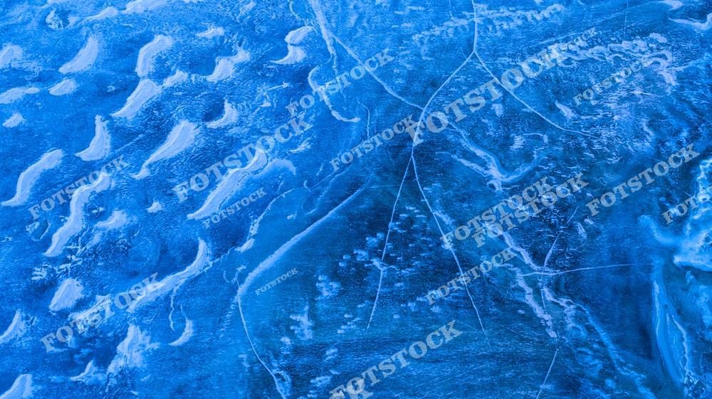 Фотография замерзшей водной поверхности, покрытой ледяными узорами и трещинами. Снимок выполнен с высоты, подчеркивая текстуры и глубину природных деталей. Идеально для иллюстрации зимней природы и замерзших водоемов.