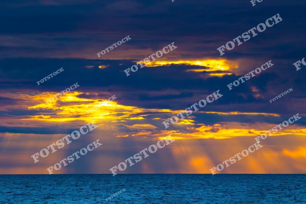 Фотография закатного неба над морем с прорывающимися через плотные облака солнечными лучами. Желто-оранжевые оттенки заката создают красивый контраст с темными облаками и спокойной поверхностью воды. Изображение передает атмосферу умиротворения и величия морского пейзажа на закате.
