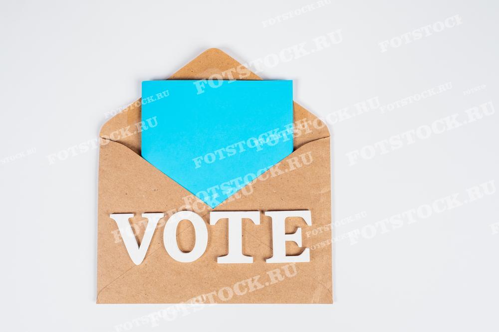Фотография демонстрирует конверт с текстом "Vote" и вставленной синей карточкой, символизирующей процесс голосования по почте.