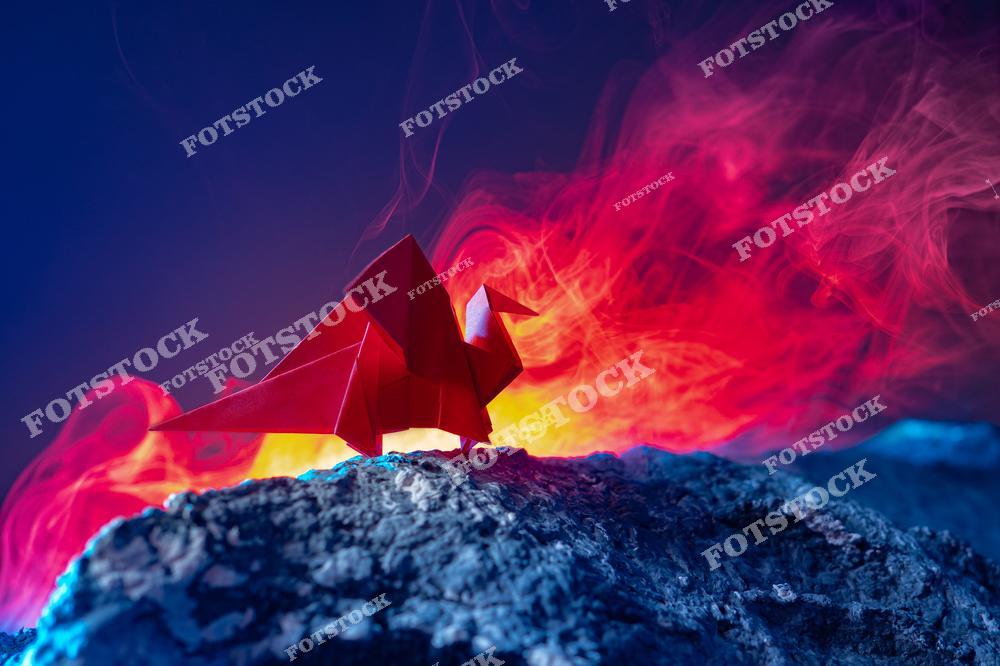 На фотографии изображен красный бумажный дракон в технике оригами, стоящий на каменистой поверхности на фоне насыщенной красной и оранжевой дымки, имитирующей пламя. Контрастные цвета и дымчатый фон создают атмосферу магии и приключений, добавляя динамику и энергию сцене. Это изображение идеально подойдет для тем, связанных с фэнтези, креативным искусством и декоративными элементами.