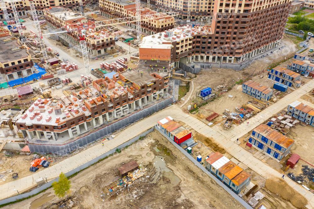 На фотографии изображена стройка жилого комплекса: несколько многоквартирных домов на разных стадиях строительства. Можно видеть краны, строительные материалы, частично возведенные здания и временные постройки для рабочих. Фото сделано с высоты, вероятно, с помощью дрона. Такое изображение может быть использовано в статьях или презентациях, посвященных градостроительству, архитектуре, строительной индустрии или в новостных материалах, рассказывающих о развитии инфраструктуры в городах.