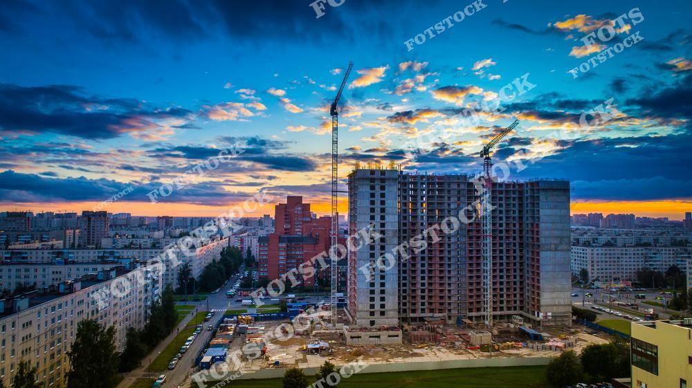 Фотография показывает строящийся многоэтажный жилой дом с башенными кранами. Вокруг расположены жилые кварталы, дороги и городская инфраструктура. Снимок иллюстрирует процесс современного городского строительства в динамично развивающемся районе.