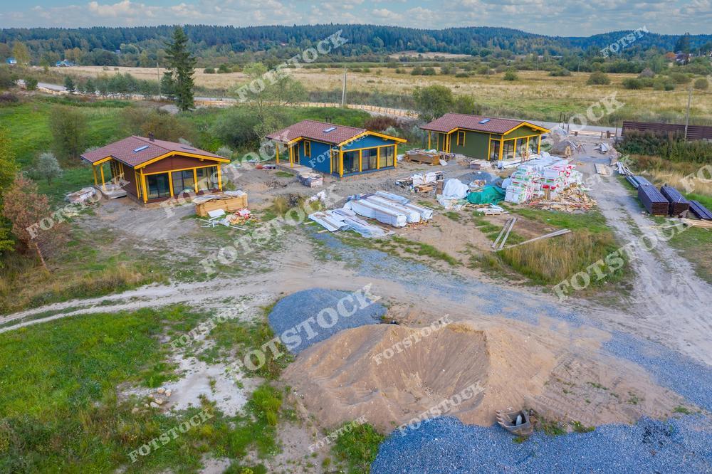 На фотографии изображены несколько загородных домов на этапе строительства в зеленой местности. На участке видны строительные материалы, техника и заготовки для дальнейших работ. Снимок подчеркивает гармонию природы и современных технологий строительства.