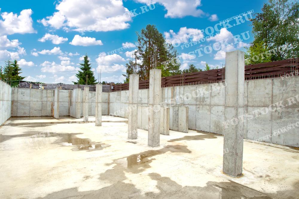 На фотографии изображен фундамент дома на начальной стадии строительства. Бетонные стены и колонны уже установлены, готовя основу для будущего здания. Видна площадка с бетонным полом, а колонны поднимаются вверх, обеспечивая поддержку для будущих конструкций. На заднем плане виден забор и зеленые деревья, что создает контраст с серыми бетонными элементами. Фотография подчеркивает этапы строительства и важность надежного фундамента для дома.