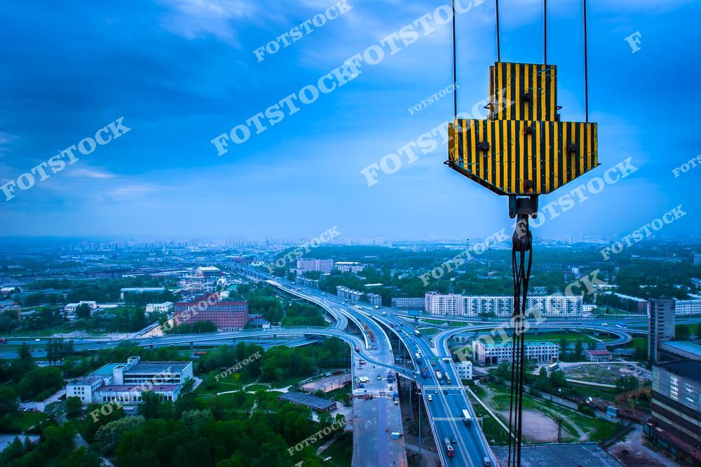 На фотографии изображен крупный строительный кран с видом на городскую инфраструктуру, включая дороги и жилые здания. Снимок сделан с высоты, показывая разветвленные дорожные развязки и панораму города на фоне вечернего неба. Фотография отражает масштабное строительство и развитие городской среды.