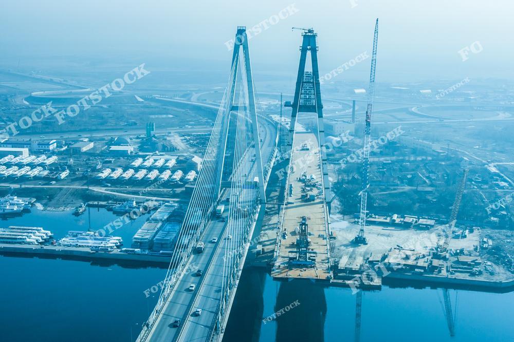 Фотография с воздуха показывает строительство вантового моста через водоем. Одна часть моста уже введена в эксплуатацию, по ней движется транспорт, а рядом продолжается возведение нового пролета с использованием подъемных кранов и строительной техники. В кадре также видны портовая зона, склады, транспортные развязки и инженерные сооружения.