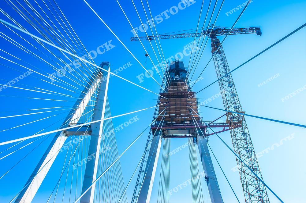 Фотография демонстрирует процесс строительства современного вантового моста с акцентом на систему тросов и строительную платформу. Вид снизу вверх показывает конструкцию моста, строительный кран и временные леса. В кадре яркое небо и архитектурные элементы будущего транспортного объекта.