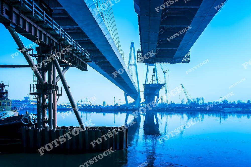 На фотографии изображена строительная конструкция вантового моста, пересекающего реку. Часть моста отражается в спокойной воде, создавая эффект глубины. Снимок подчеркивает сложность инженерных решений и значимость развития транспортной инфраструктуры.