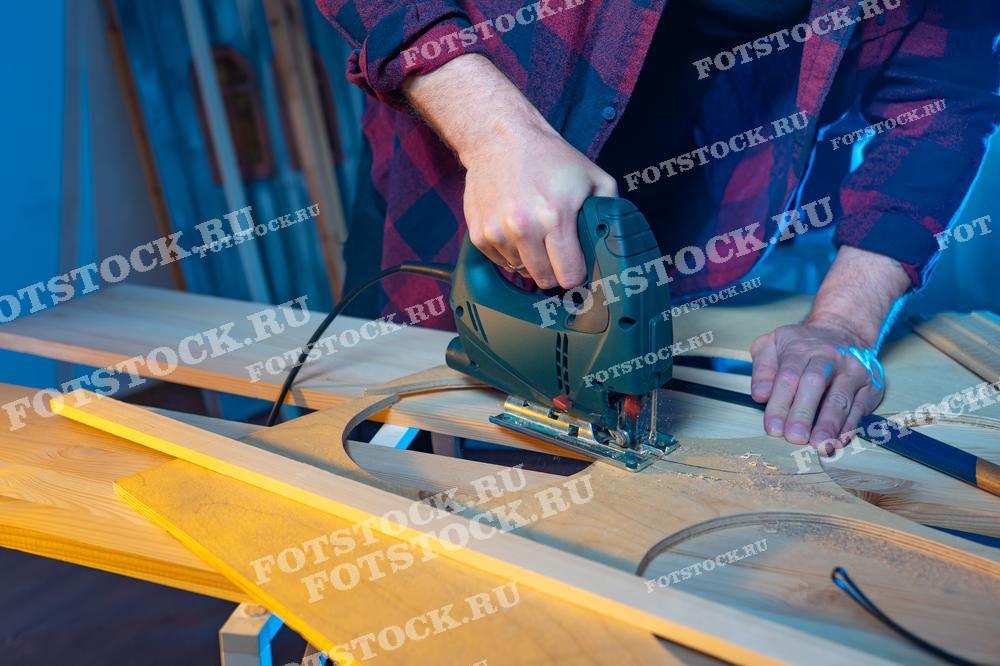 На фото изображен мастер, который с помощью электрического лобзика вырезает детали из деревянной доски. Это демонстрирует процесс деревообработки в мастерской, где важна точность и аккуратность при работе с инструментами. Фотография подчеркивает мастерство и внимание к деталям, необходимые для создания качественных элементов для строительства или ремонта.