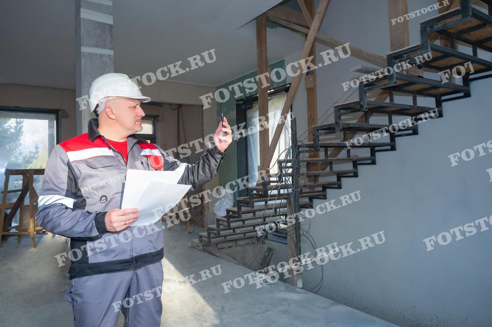 На фото изображен инженер в каске и рабочей одежде, который находится на стройплощадке. Он держит в руках документы и, вероятно, оценивает или фиксирует прогресс строительных работ, используя смартфон или другое устройство. На заднем плане видна частично построенная лестница, указывающая на стадии строительства или ремонта.