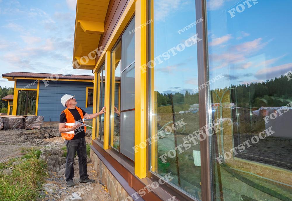 На фотографии изображен строитель в защитной каске и оранжевом жилете, осматривающий окна дома. Фасад здания выполнен из желтых и коричневых панелей с большими стеклянными окнами. На заднем плане виден еще один строящийся дом и природный ландшафт.