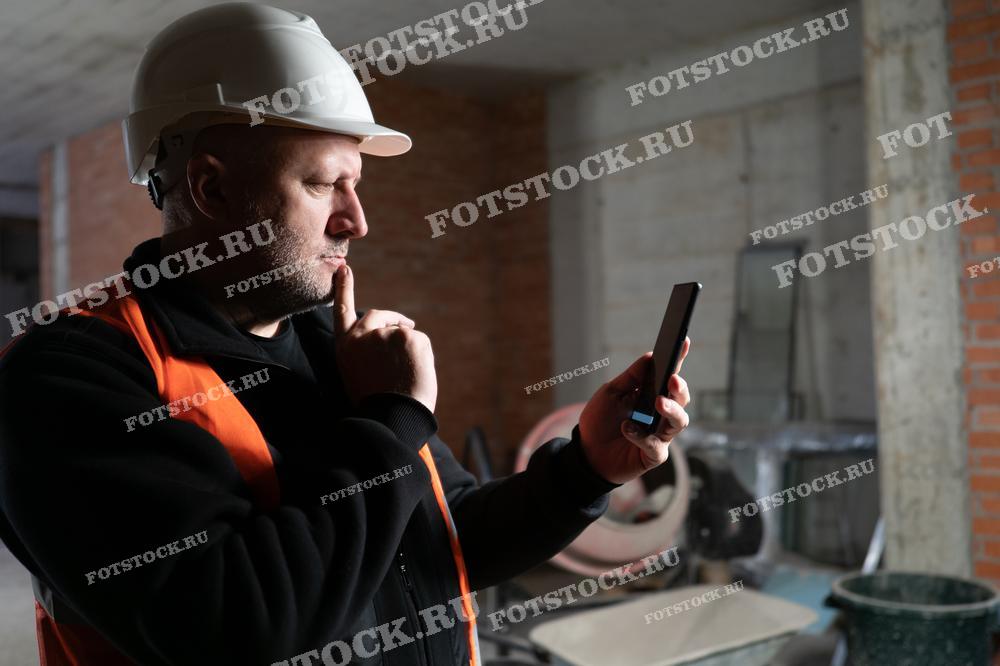 На фотографии изображен инженер в защитной каске и жилете, проверяющий информацию на смартфоне или созванивающийся с заказчиком. Он находится на строительной площадке внутри здания, что демонстрирует процесс контроля качества и выполнения внутренних строительных работ.