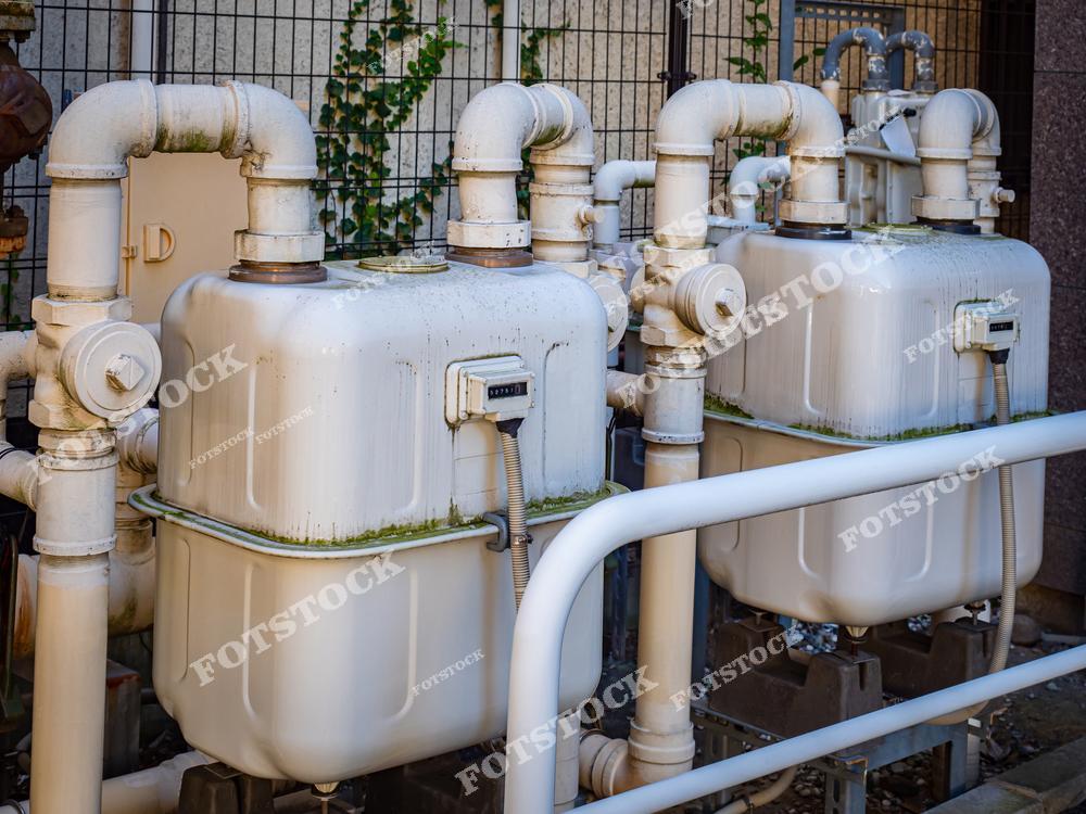 На фотографии изображены уличные газовые счетчики с подключенными к ним трубами и коммуникациями. Оборудование установлено возле здания и огорожено защитной конструкцией. Фотография иллюстрирует элементы инженерных систем для учета и распределения газа.