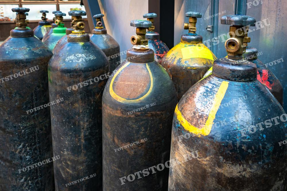 На фотографии изображены газовые баллоны, используемые для сварочных работ, стоящие в промышленном помещении. Баллоны различных цветов и размеров предназначены для хранения и транспортировки сварочного газа, необходимого для выполнения различных видов сварочных процессов.