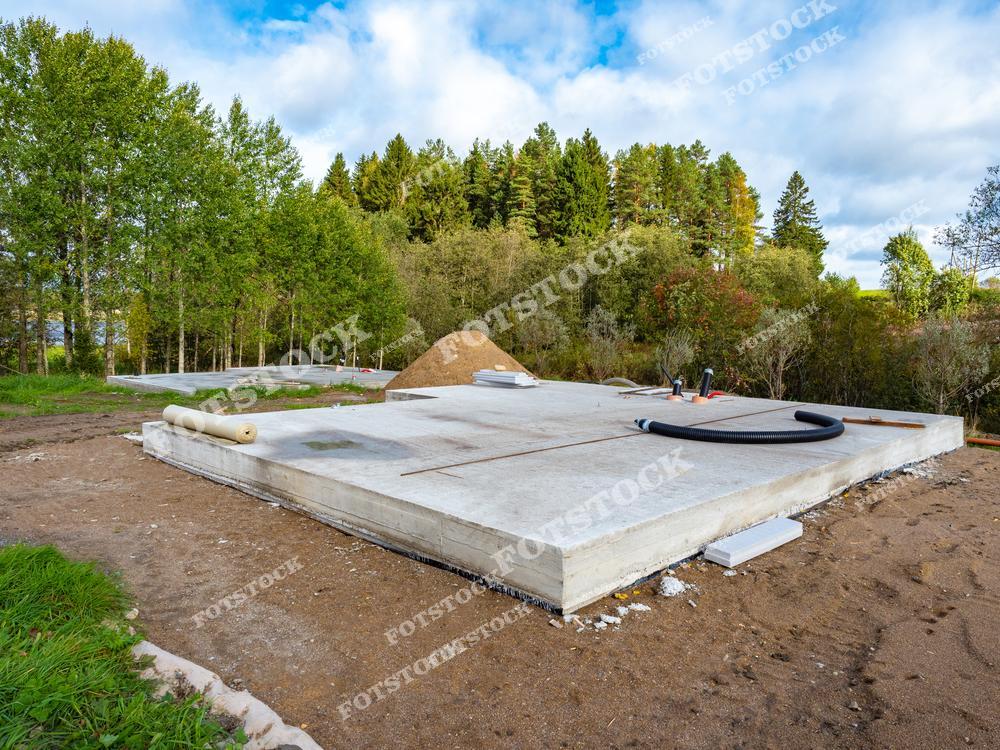 На фотографии показан бетонный фундамент для загородного дома, окружённый зелёным лесом. Видны подготовительные работы и инженерные коммуникации. Снимок символизирует начальный этап строительства и выбор экологичного места для жилья.