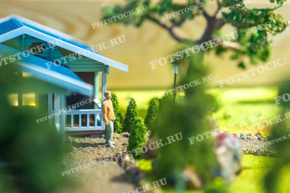 На фотографии изображен макет деревянного дома с голубой крышей и фигурка человека рядом с ним. Макет окружен миниатюрными деревьями и кустарниками, создавая ощущение загородного участка. Фото может быть использовано для иллюстрации материалов по архитектуре и проектированию, статьях о ландшафтном дизайне и строительстве, а также в рекламных материалах компаний, занимающихся строительством домов и ландшафтным дизайном.