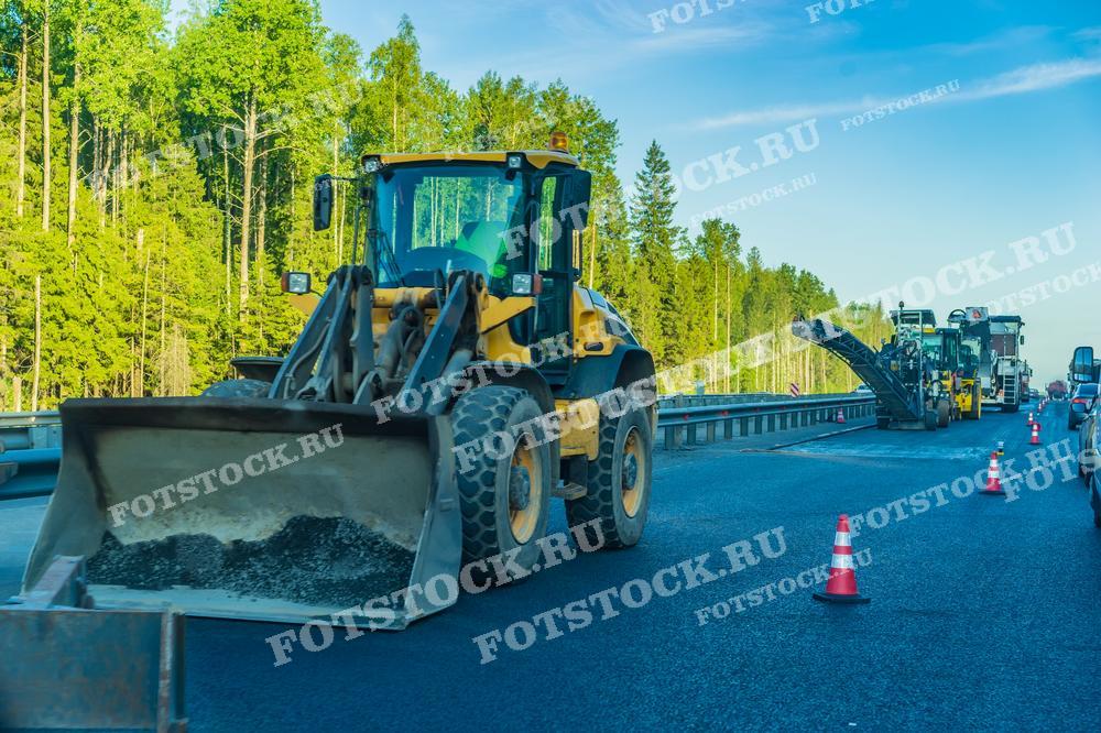 На фотографии изображен процесс дорожных работ на трассе. В кадре видны несколько строительных машин, включая погрузчик и асфальтоукладчики. Дорога огорожена дорожными конусами для безопасности. Работы проводятся на фоне зеленого леса, что создает контраст между природой и техникой. Этот снимок может быть использован в статьях о дорожном строительстве, в образовательных материалах по гражданскому строительству, а также в рекламных материалах компаний, занимающихся дорожными работами.