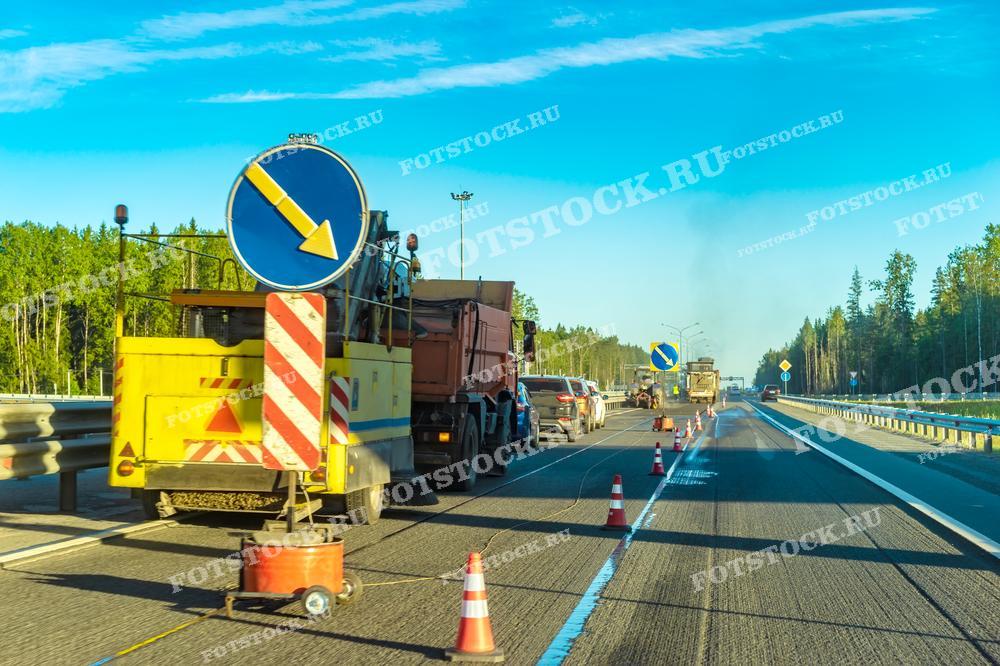 На фото изображены дорожные работы на автомагистрали, где временно изменено направление движения с помощью дорожных знаков и конусов. Видна специализированная техника и рабочие, занимающиеся ремонтом и обслуживанием дорожного покрытия. На заднем плане — лесная зона, создающая контраст с оживленной магистралью.