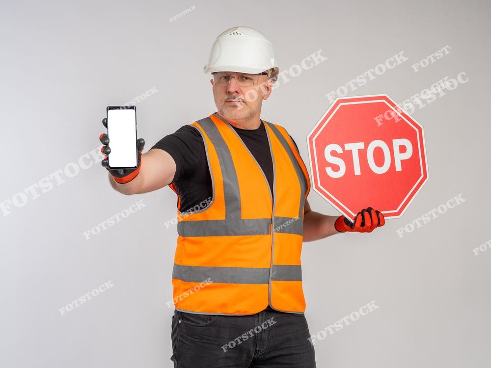 На фотографии изображен рабочий в каске и жилете, держащий табличку «STOP» и смартфон с пустым экраном. Это подчеркивает важность безопасности и современных технологий в строительной сфере. Снимок символизирует ответственность и контроль на объектах.