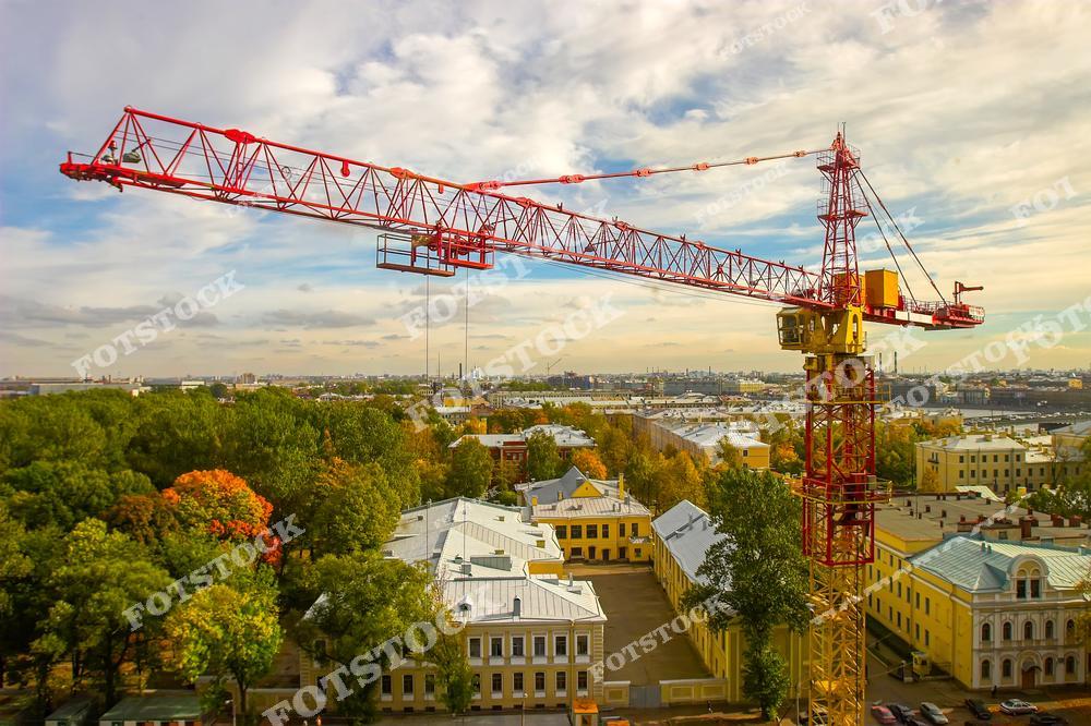Красно-жёлтый башенный кран возвышается над городскими кварталами с зелёными парками и старинными зданиями. Вдалеке виден горизонт с промышленными объектами. Фото передаёт процесс строительства и реконструкции в городской среде.