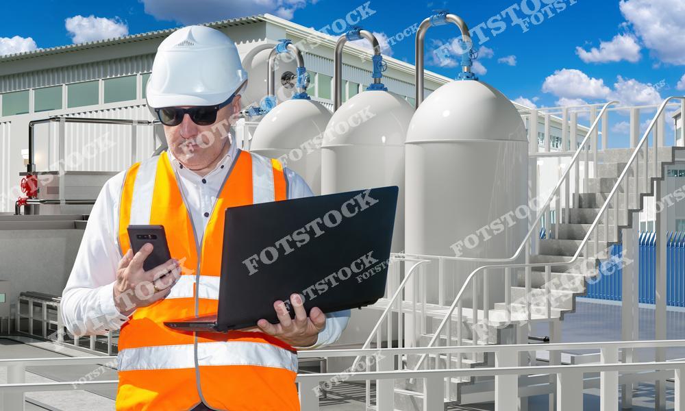 Мужчина-инспектор в рабочей униформе и защитной каске стоит рядом с промышленными резервуарами на нефтехимическом заводе. В одной руке он держит телефон, а в другой — ноутбук, что указывает на его работу с технологическими данными и мониторинг состояния оборудования. Этот кадр подчеркивает важность экологического контроля и безопасности на крупных промышленных объектах.