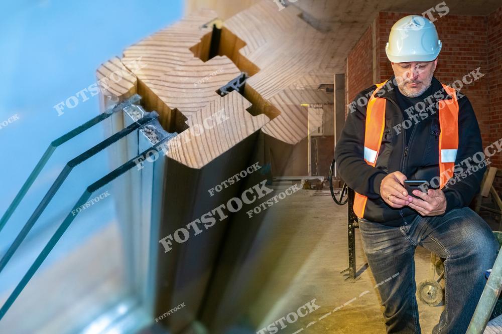 На фотографии изображены крупным планом конструкция деревянного стеклопакета и инженер на строительной площадке. Инженер в каске и жилете использует смартфон для работы. Снимок демонстрирует технологические решения и процесс проверки качества в строительстве.
