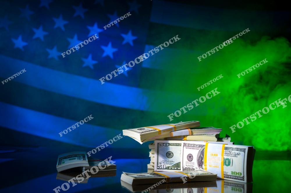 На изображении представлены аккуратно уложенные пачки долларов США различных номиналов на фоне стилизованного американского флага и зелёного дыма. Фото символизирует финансовую силу, экономику, богатство и влияние США. Подходит для тем, связанных с финансами, инвестициями, банковской системой и международной экономикой.