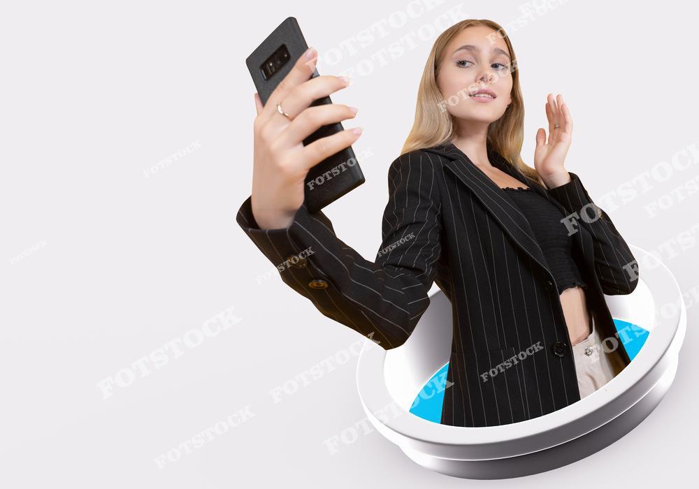 На изображении молодая женщина в стильном деловом костюме держит смартфон, позируя для селфи. Её уверенный взгляд и жест подчеркивают современную тему самопрезентации в интернете. Снимок символизирует активное использование социальных сетей и мобильных устройств.