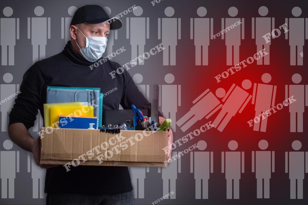 На изображении мужчина в защитной маске держит коробку с офисными вещами. Фон состоит из силуэтов людей, среди которых выделены красным падающие фигуры, что подчеркивает тему увольнений и экономического спада. Атмосфера напряженная и мрачная, подчеркнутая серым и красным цветами изображения.