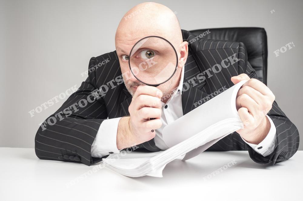 На изображении мужчина в деловом костюме держит лупу, через которую смотрит на большой стопку документов. Его выражение лица передает сосредоточенность, символизируя процесс анализа и поиска деталей. Иллюстрация офисной работы, проверки и контроля.
