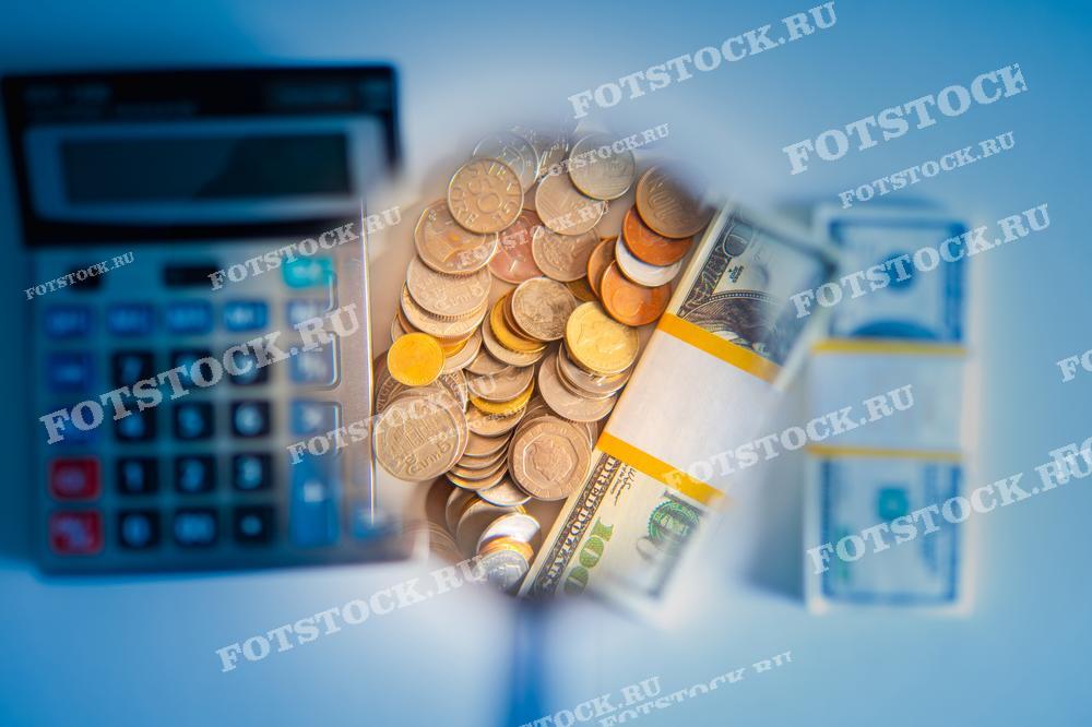 Фотография показывает монеты и банкноты под увеличительным стеклом на фоне калькулятора. Это изображение символизирует финансовый анализ, управление бюджетом и бухгалтерию.