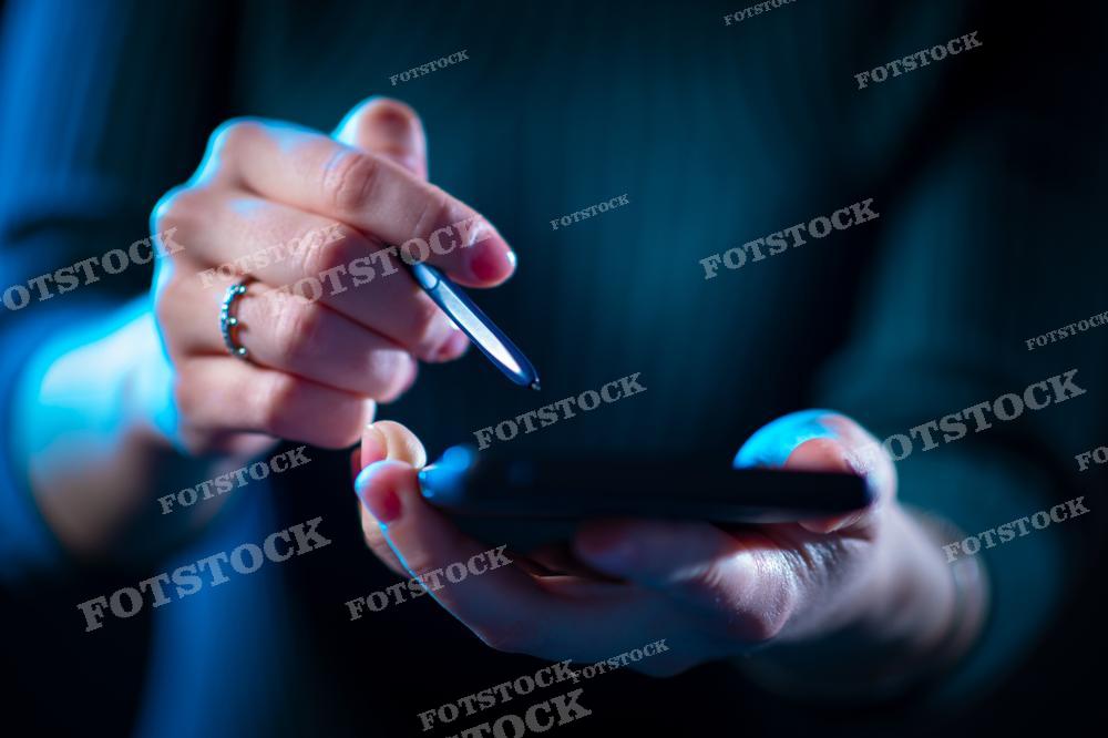а фотографии крупным планом показаны руки человека, работающего со смартфоном. Одна рука держит устройство, в то время как другая использует стилус для навигации по экрану. Синеватый свет и фокусировка на руках создают современное и технологичное настроение. Этот снимок может быть использован для иллюстрации материалов, связанных с мобильными технологиями, цифровой коммуникацией или бизнес-приложениями.