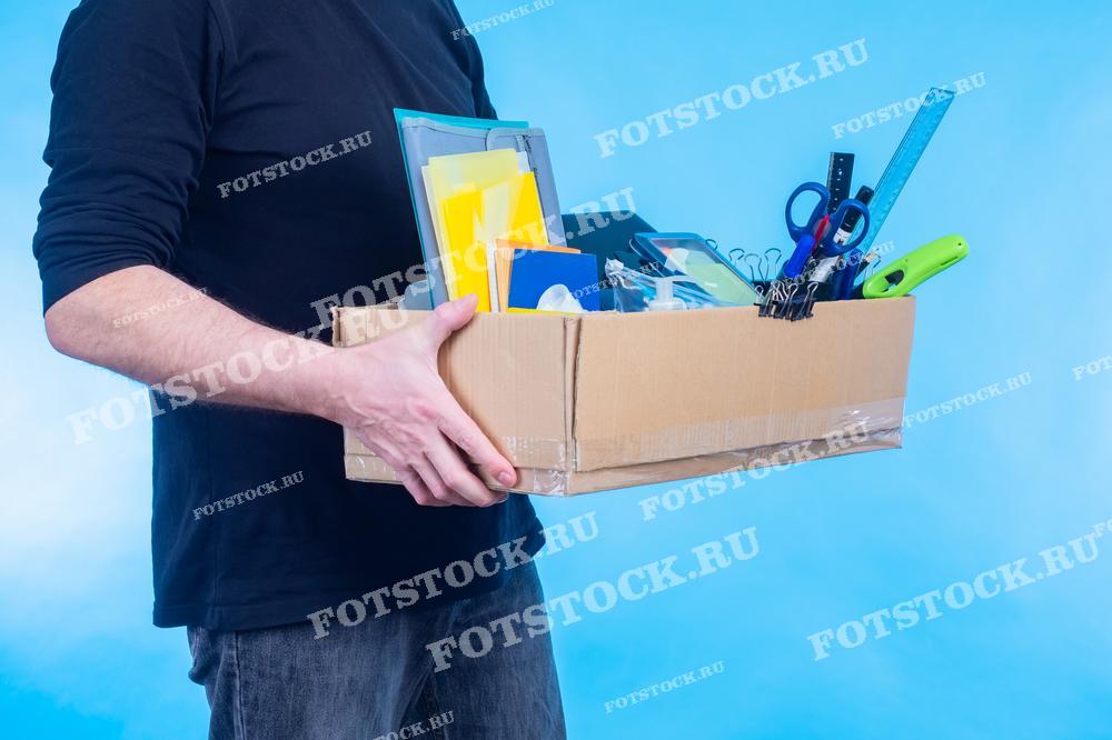 Фотография показывает человека, держащего коробку с офисными принадлежностями, такими как папки, ручки, линейки и скрепки, на фоне синего цвета. Это изображение символизирует процесс увольнения, смены рабочего места или перемещения в новый офис.
