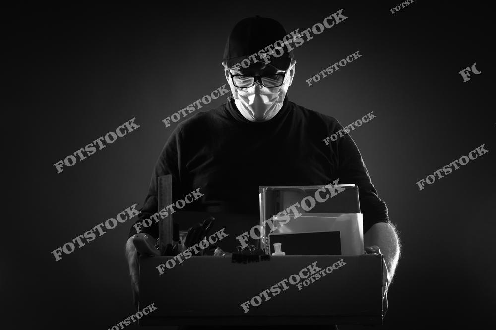 На черно-белой фотографии человек в медицинской маске стоит у закрытой фирмы с коробкой в руках, символизируя увольнение и потерю работы из-за эпидемии коронавируса. Это изображение отражает экономический кризис, массовые сокращения штатов и закрытие компаний, вызванные пандемией.