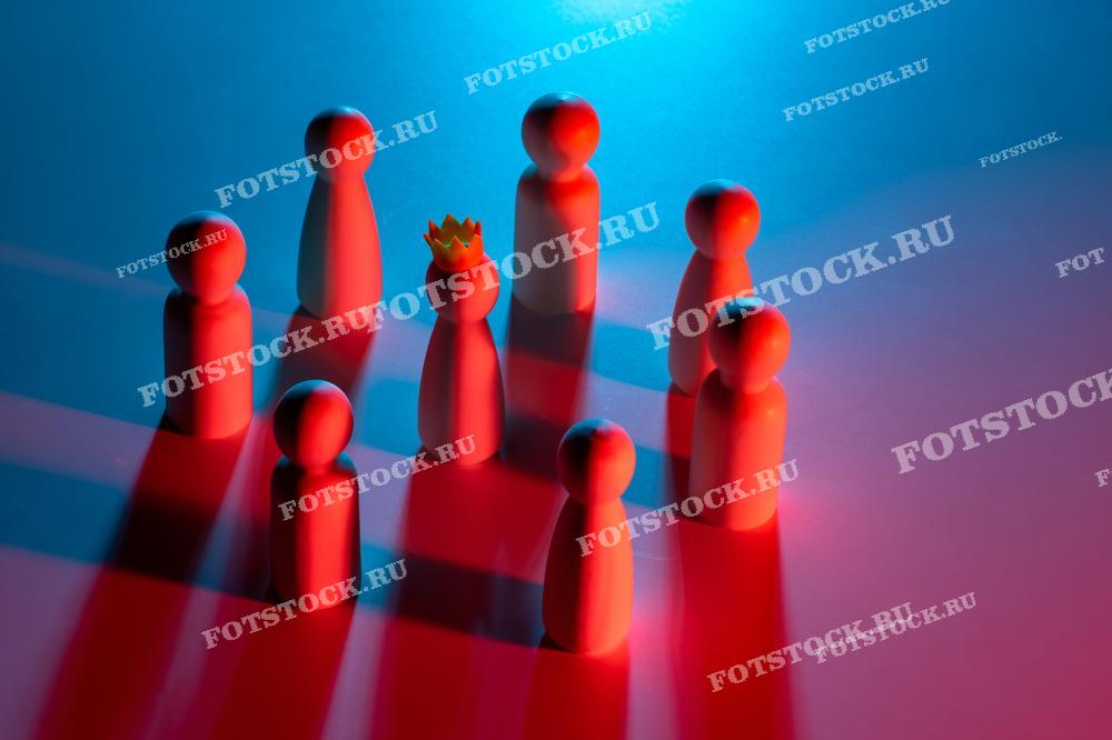 Фотография показывает группу деревянных фигурок, освещенных синим и красным светом, одна из которых выделяется короной. Это изображение символизирует лидерство, командную работу и иерархию.