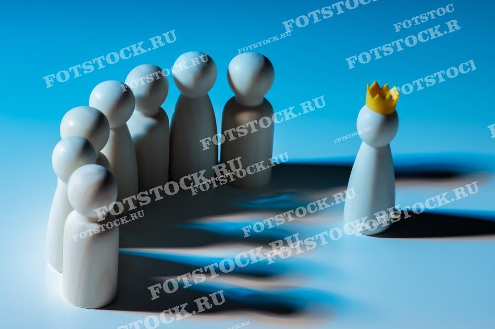 Фотография показывает группу деревянных фигурок, стоящих перед одной фигуркой с короной на синем фоне. Это изображение символизирует лидерство, командную работу и иерархию.