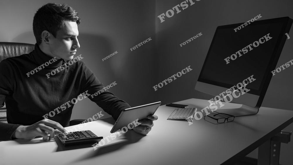 На фотографии изображен мужчина в офисной обстановке, использующий планшет и калькулятор. Рядом с ним находятся компьютер и очки, создавая атмосферу сосредоточенной работы. Черно-белая палитра подчёркивает деловую обстановку и акцент на аналитике.