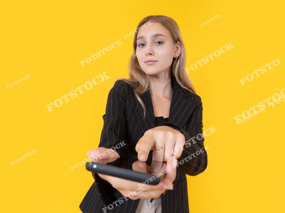 На фотографии изображена девушка, которая протягивает смартфон и указывает на его экран. Она выглядит уверенно, а желтый фон подчеркивает динамичность и яркость ситуации. Снимок символизирует использование технологий в повседневной жизни.
