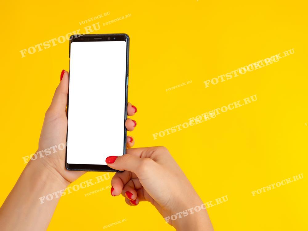 Фотография показывает женскую руку с красным маникюром, держащую смартфон с пустым белым экраном на ярко-желтом фоне. Это изображение символизирует использование современных технологий и гаджетов в повседневной жизни.