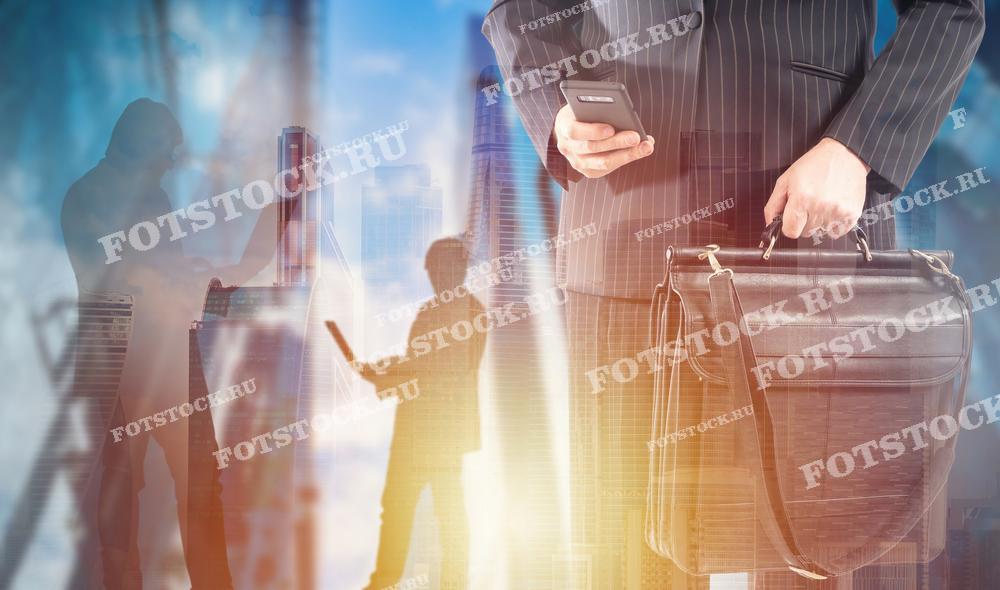 Фотография показывает бизнесмена в костюме, держащего телефон и портфель на фоне городской застройки и силуэтов других деловых людей. Это изображение символизирует деловую активность, современный бизнес и коммуникации.