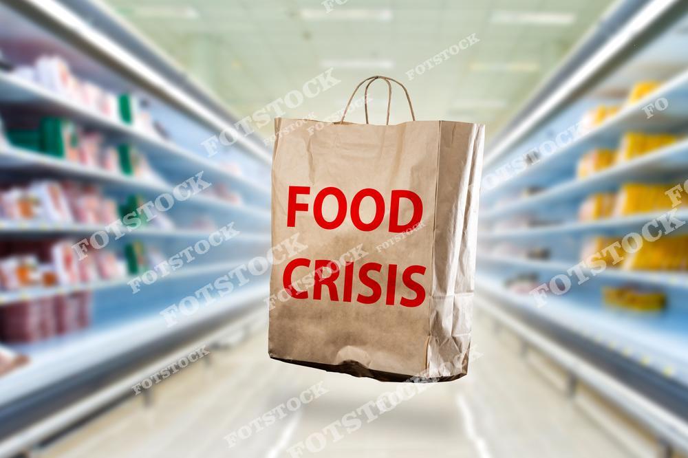 отография показывает бумажный пакет с надписью «Food Crisis» на фоне размытого изображения полок в магазине. Изображение отражает проблему глобального продовольственного кризиса и нехватки продуктов. Символический образ нестабильности и неопределенности в обеспечении продовольствием.