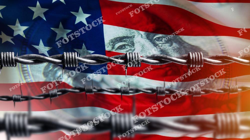 На изображении фрагмент флага США и купюра с портретом Бенджамина Франклина, пересеченные колючей проволокой. Композиция выражает идею препятствий, ограничений и экономического давления.