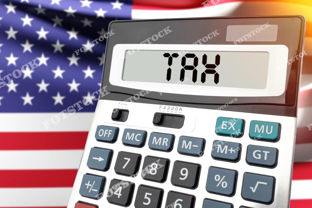 На переднем плане изображен калькулятор с надписью "Tax" на экране. На заднем плане — развевающийся флаг США, подчеркивающий национальный контекст налоговых расчетов.